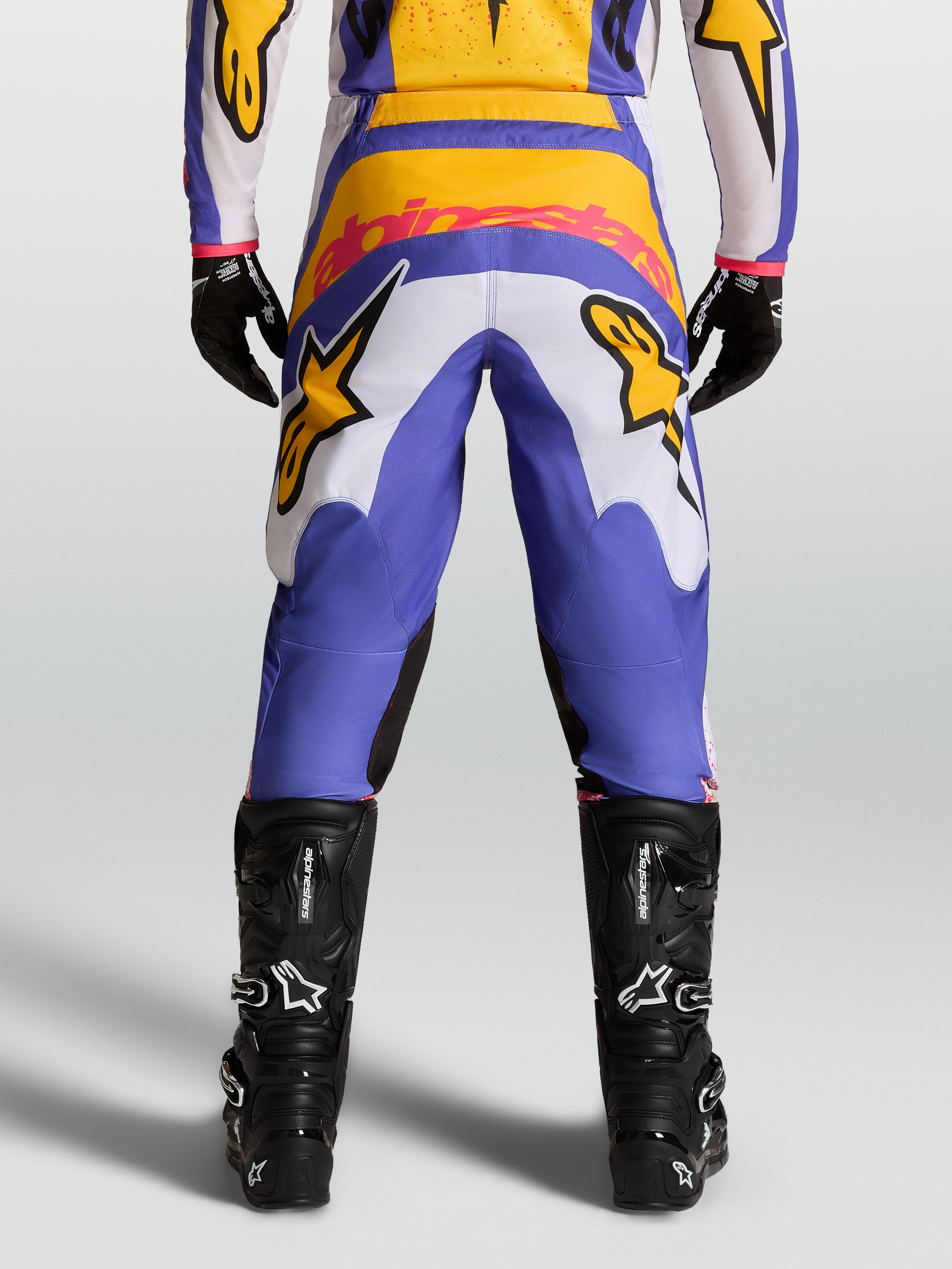 Alpinestars Fluid Drag Pants for motocross racing, view from behind, in Blue and Gold with stretch corduroy panels and reinforced seat, paired with black technical boots.