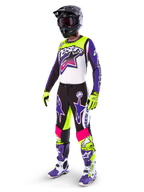 Motocyklista motocrossowy w stroju Alpinestars Limited Edition Supertech Dirt Studios®, kolorystyka biało-fioletowa i żółty fluo, czarne spodnie z wyrazistymi fioletowymi i różowymi logotypami gwiazdy, neonowożółte rękawice, fioletowo-białe buty wyścigowe, czarny kask, kompletny profesjonalny zestaw wyścigowy do jazdy terenowej