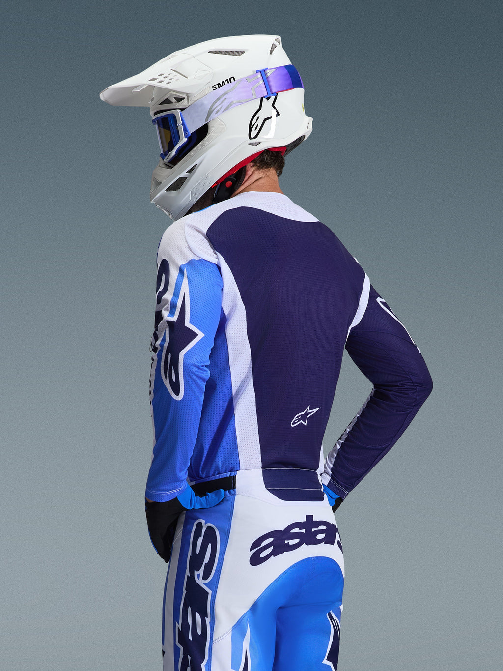 Motocyklista motocrossowy w koszulce Alpinestars 2026 Racer Air Portl w kolorze Ucla Blue i białym, widok z tyłu eksponujący oddychający granatowy panel siatkowy na plecach, zestawiony z dopasowanymi spodniami i białym kaskiem Alpinestars z niebieskimi goglami, spójny komplet odzieży motocrossowej.