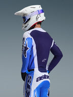 Motocyklista motocrossowy w koszulce Alpinestars 2026 Racer Air Portl w kolorze Ucla Blue i białym, widok z tyłu eksponujący oddychający granatowy panel siatkowy na plecach, zestawiony z dopasowanymi spodniami i białym kaskiem Alpinestars z niebieskimi goglami, spójny komplet odzieży motocrossowej.