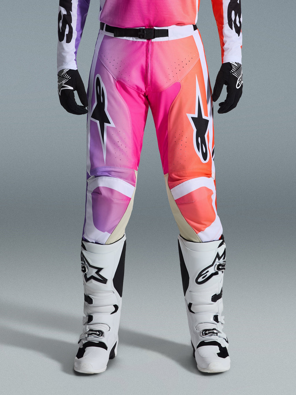Alpinestars 2026 Racer Air Portl Pants, spodnie motocrossowe, gradient w intensywnych odcieniach fioletu, różu i pomarańczu, z wyraźnymi logotypami gwiazdy na udach, zestawione z białymi butami wyścigowymi i czarnymi rękawicami, zaprojektowane do wyczynowej jazdy terenowej