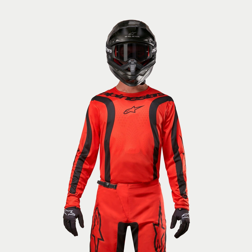 Zawodnik motocrossowy w koszulce Alpinestars 2024 Fluid Lurv, jaskrawopomarańczowa i czarna, pasujące spodnie motocrossowe, czarny kask z goglami i czarne rękawice, skoordynowany zestaw do wyścigów terenowych