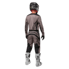 Alpinestars 2024 Fluid Lurv Jersey, koszulka motocrossowa, Mud Black, widok z tyłu zawodnika motocrossowego w dopasowanym stroju wyścigowym, czarny kask i białe buty, wyraźne czarne logotypy Alpinestars, sportowy krój zapewniający wydajność w terenie