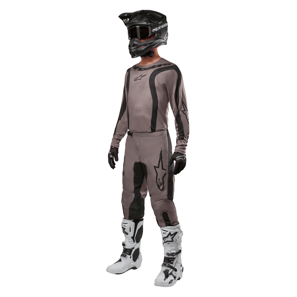 Zawodnik motocrossowy ubrany w pełny strój wyścigowy z koszulką Alpinestars 2024 Fluid Lurv w kolorze Mud Black, w zestawieniu z dopasowanymi spodniami, czarnym kaskiem z goglami, biało-czarnymi butami i czarnymi rękawicami, skoordynowany zestaw do wyścigów terenowych