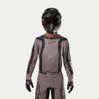 Motocyklista motocrossowy w koszulce Alpinestars 2024 Fluid Lurv w kolorze Mud Black, wykonanej z ziemistej szarobrązowej tkaniny z czarnymi graficznymi pasami i logo gwiazdy na piersi, w połączeniu z czarnym kaskiem, czerwonymi goglami i czarnymi rękawicami dla spójnego wyścigowego wyglądu off-road
