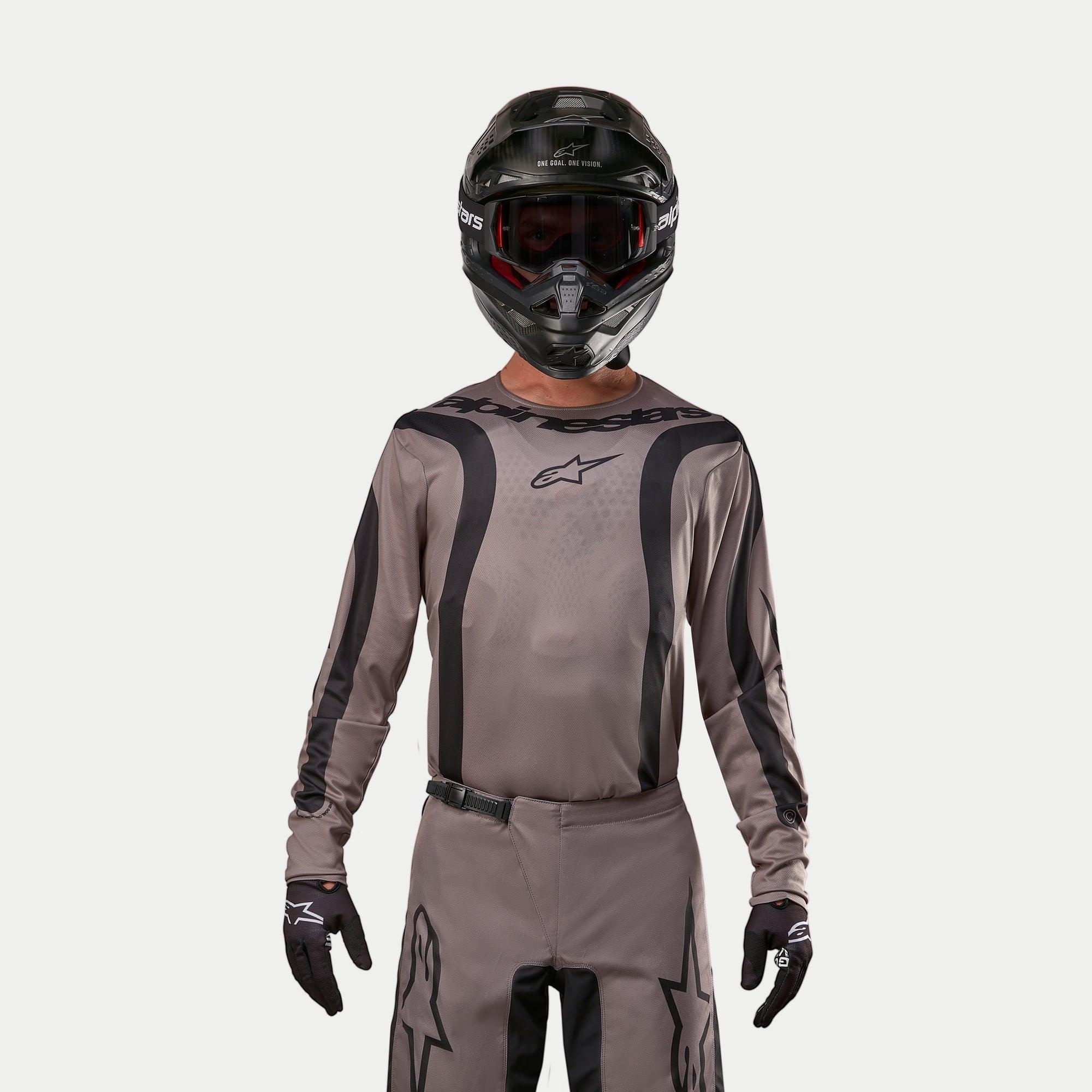 Motocyklista motocrossowy w koszulce Alpinestars 2024 Fluid Lurv w kolorze Mud Black, wykonanej z ziemistej szarobrązowej tkaniny z czarnymi graficznymi pasami i logo gwiazdy na piersi, w połączeniu z czarnym kaskiem, czerwonymi goglami i czarnymi rękawicami dla spójnego wyścigowego wyglądu off-road