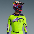 Zawodnik motocrossowy w stroju Alpinestars Supertech Vista 2026, neonowożółta koszulka z fioletowymi i czarnymi detalami, duże fioletowe logo z gradientem na piersi, wielokolorowy kask i fioletowe gogle, laserowo wycinana wentylacja, profesjonalny sportowy krój