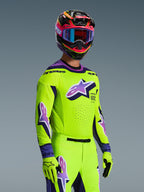 Zawodnik motocrossowy w koszulce Alpinestars 2026 Supertech Vista, jaskrawożółta z fioletowymi i czarnymi akcentami, pasujące żółte spodnie i rękawice, wielokolorowy kask z fioletowymi goglami, laserowo wycinane perforacje i duże logo Astar na piersi, profesjonalny strój wyścigowy