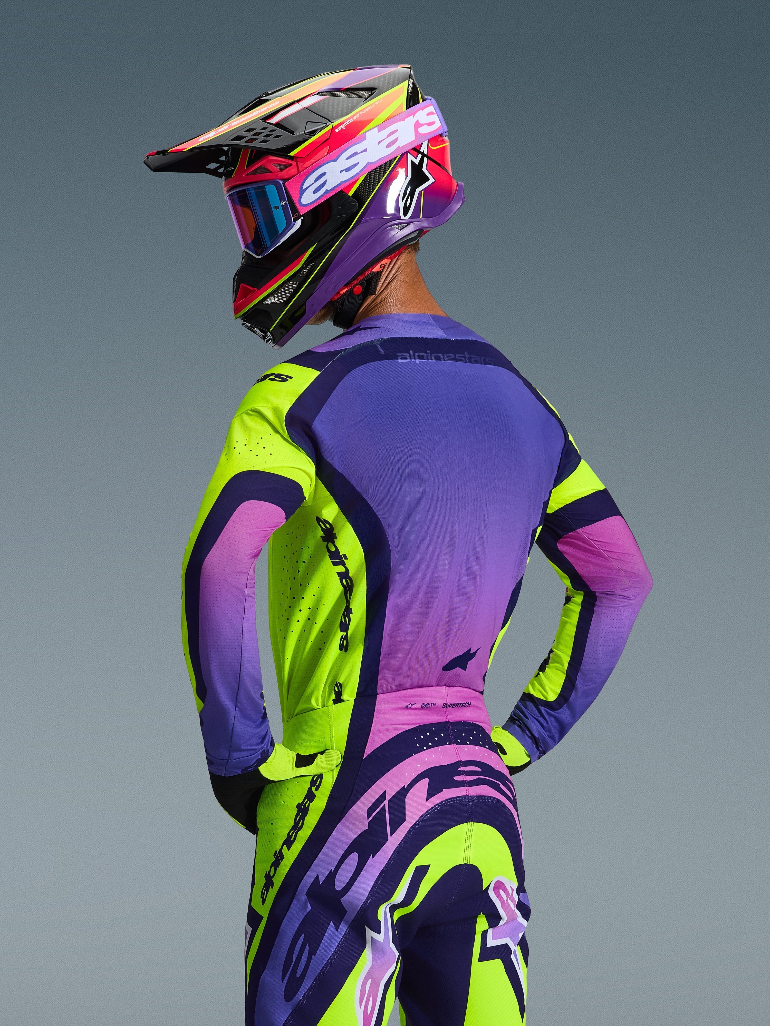 Zawodnik motocrossowy widziany od tyłu w koszulce Alpinestars Supertech Vista 2026 i pasujących spodniach, kolorystyka Yellow Purple Black z jaskrawymi gradientami, dopasowany kask wyścigowy z niebieskimi goglami i żółtymi rękawicami, profesjonalny zestaw off-road o sportowym kroju