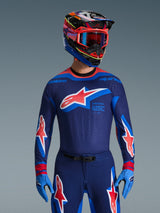 Alpinestars 2026 Supertech Vista Jersey, koszulka motocrossowa, ciemnogranatowa z koralowymi i kobaltowymi akcentami, noszona z pasującymi spodniami i kaskiem, z dużym logo gwiazdy na piersi i brandingiem race spec, sportowy krój z wentylowanymi panelami z siatki.