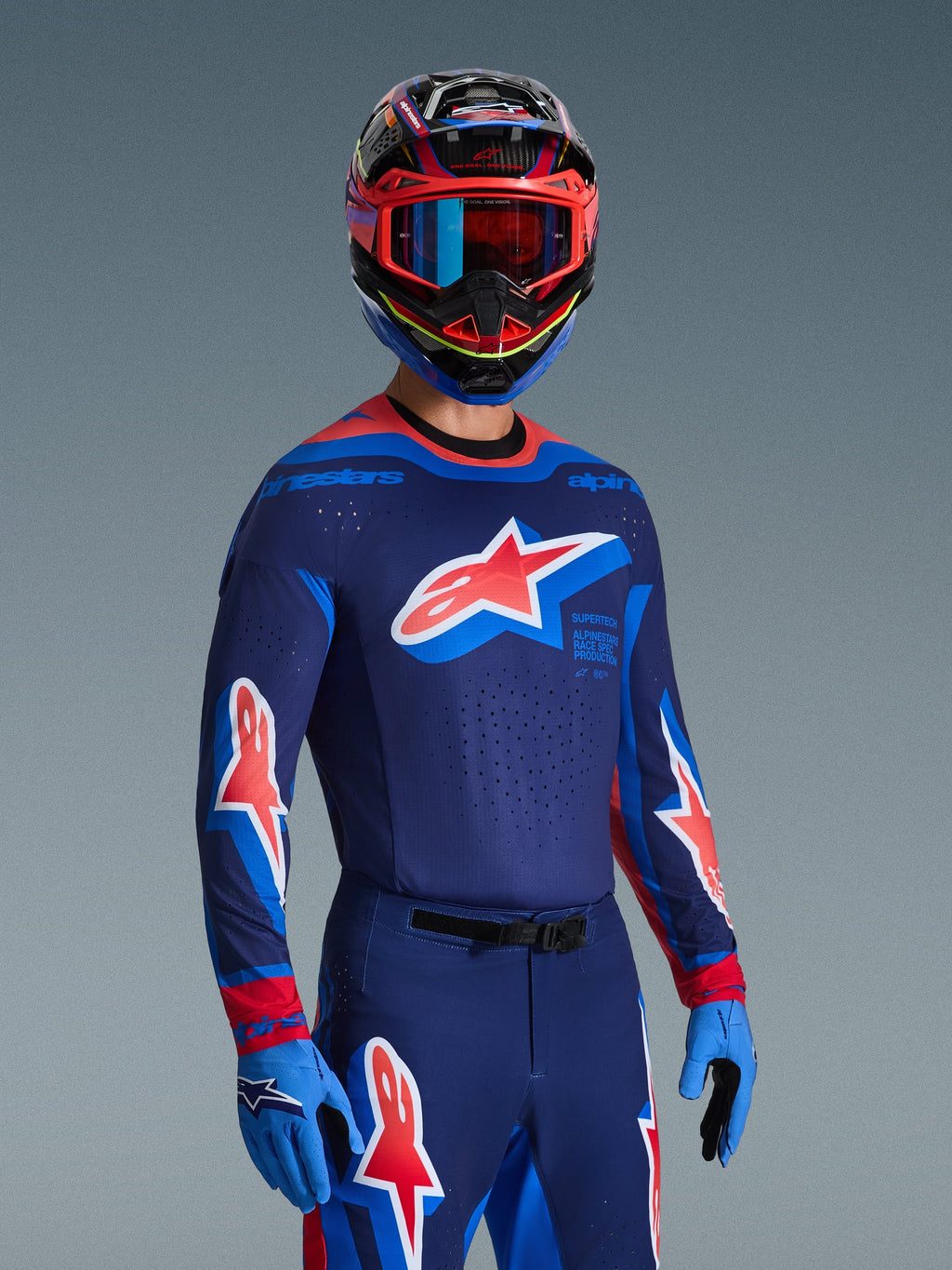 Zawodnik motocrossowy w koszulce 2026 Supertech Vista w kolorach ciemnogranatowym, koralowym i kobaltowym, pasujące spodnie i rękawice, wielokolorowy kask z czerwonymi goglami, profesjonalny zestaw odzieży do wyścigów terenowych