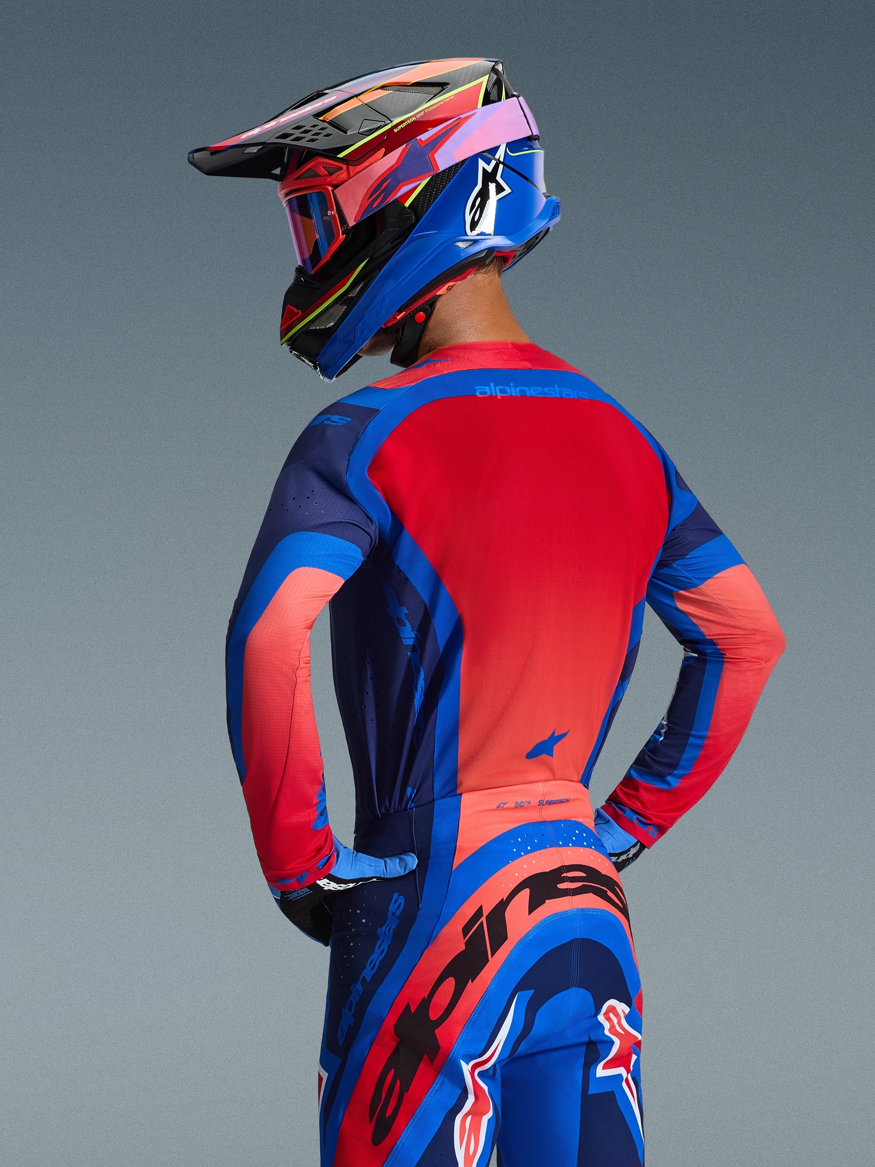 Widok z tyłu zawodnika motocrossowego ubranego w koszulkę Alpinestars 2026 Supertech Vista w kolorach Dark Navy, Hot Coral i Cobalt, z wyrazistym czerwonym panelem tylnym i bezszwową konstrukcją, w zestawie z pasującymi spodniami i kaskiem, tworzącymi kompletny wyścigowy wygląd.