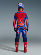 Zawodnik motocrossowy widziany od tyłu w zestawie Alpinestars 2026 Supertech Vista w kolorach granatowym, koralowym i kobaltowym, z jaskrawoczerwonym tyłem koszulki, wyraźnym brandingiem na spodniach, dopasowanymi czerwonymi butami oraz niebiesko-różowym kaskiem