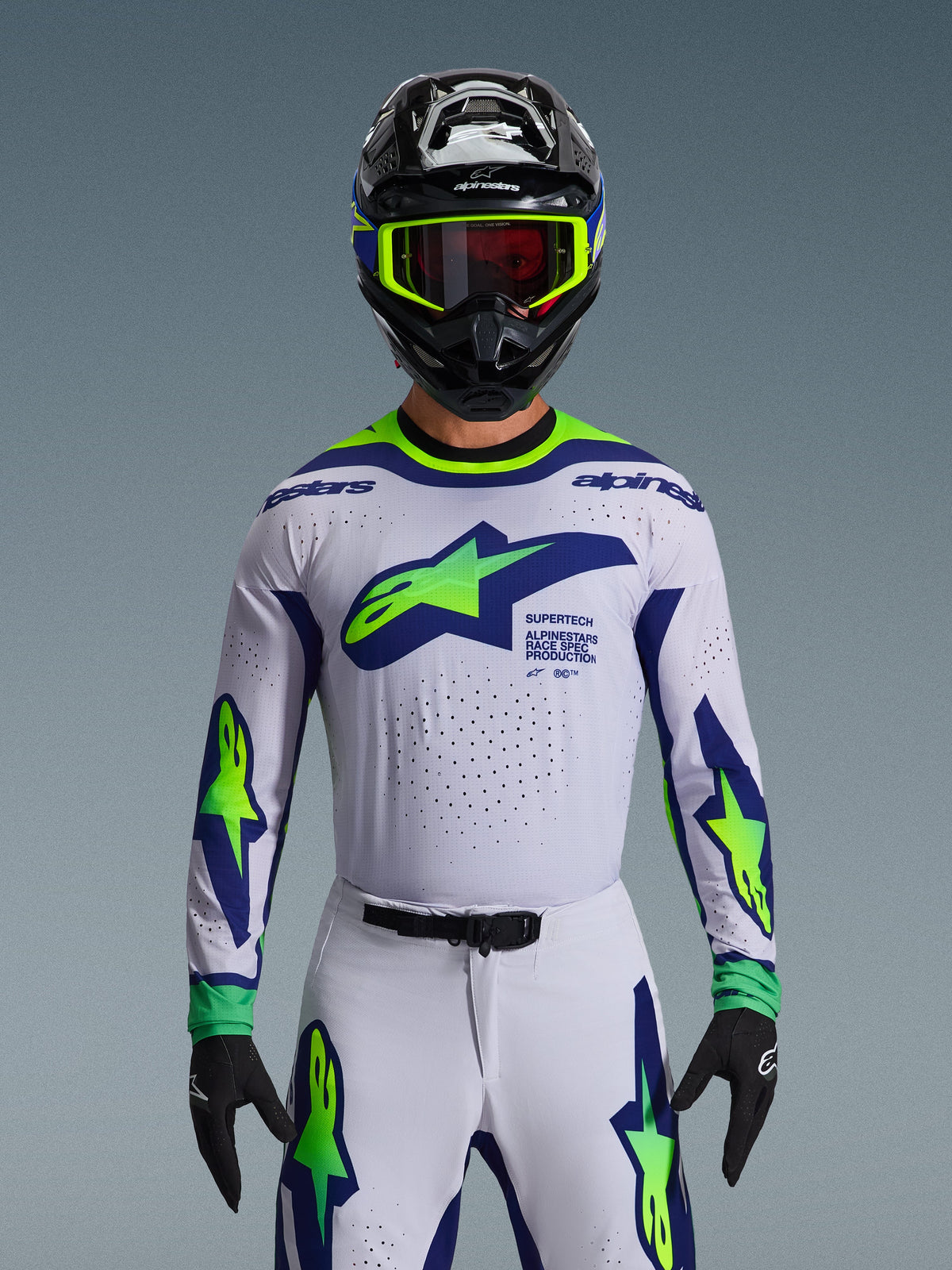 Alpinestars 2026 Supertech Vista Jersey, koszulka motocrossowa, jasnoszara, żółta fluo i niebieska, noszona przez zawodnika z pasującymi spodniami i czarnym kaskiem, duże logo na klatce piersiowej, laserowo wycinane perforacje wentylacyjne, sportowy krój pro-level