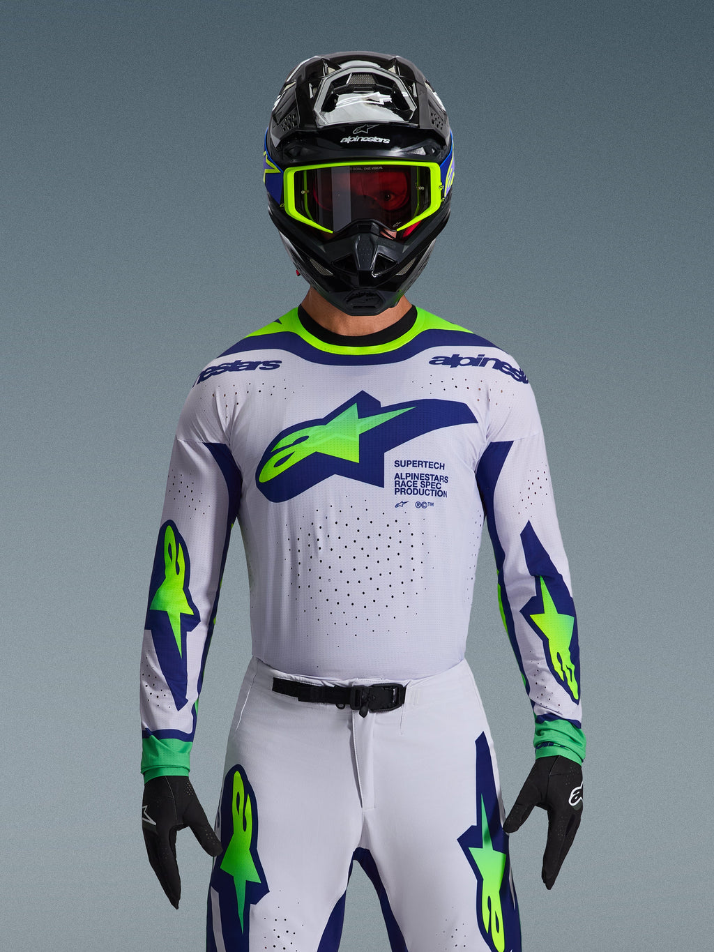 Alpinestars 2026 Supertech Vista Jersey, koszulka motocrossowa, jasnoszara, żółta fluo i niebieska, noszona przez zawodnika z pasującymi spodniami i czarnym kaskiem, duże logo na klatce piersiowej, laserowo wycinane perforacje wentylacyjne, sportowy krój pro-level