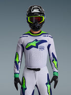 Alpinestars 2026 Supertech Vista Jersey, koszulka motocrossowa, jasnoszara, żółta fluo i niebieska, noszona przez zawodnika z pasującymi spodniami i czarnym kaskiem, duże logo na klatce piersiowej, laserowo wycinane perforacje wentylacyjne, sportowy krój pro-level
