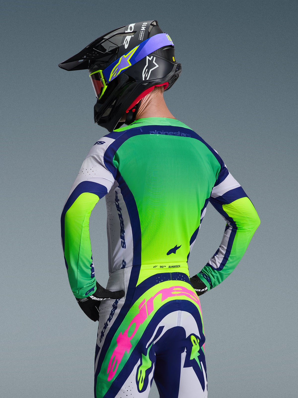 Kierowca motocrossowy widziany od tyłu, ubrany w koszulkę Alpinestars 2026 Supertech Vista w kolorystyce Light Gray Yellow Fluo Blue, w zestawie z pasującymi spodniami i kaskiem, z jaskrawymi neonowymi panelami i sportowym krojem wyścigowym do jazdy w terenie.