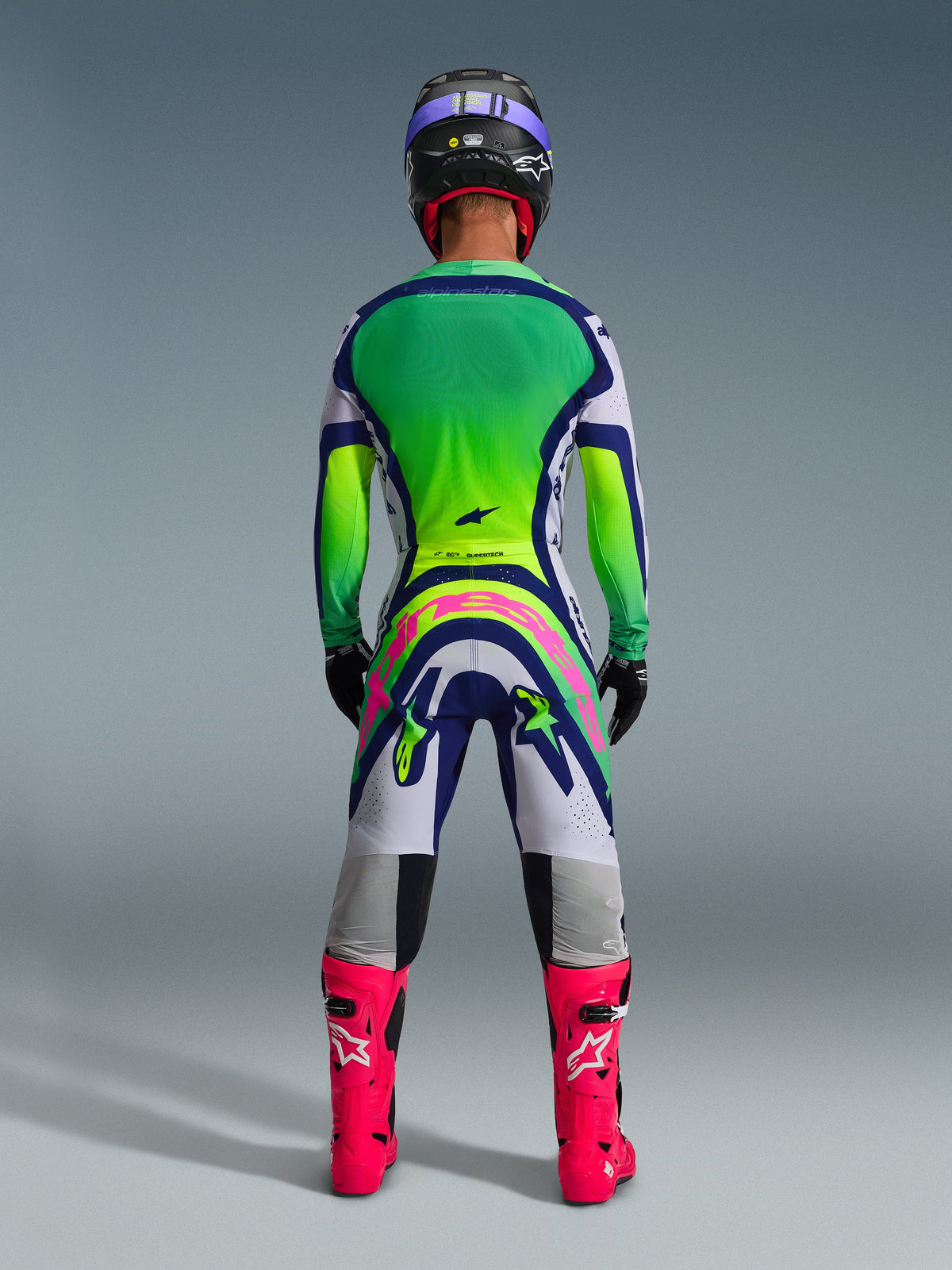 Strój motocrossowy Alpinestars, kompletny zestaw wyścigowy widziany od tyłu, koszulka Supertech Vista 2026 z zielono-żółtym gradientem na plecach, biało-niebieskie spodnie z dużym różowym logo Alpinestars, jaskraworóżowe buty oraz czarny kask z fioletowymi akcentami