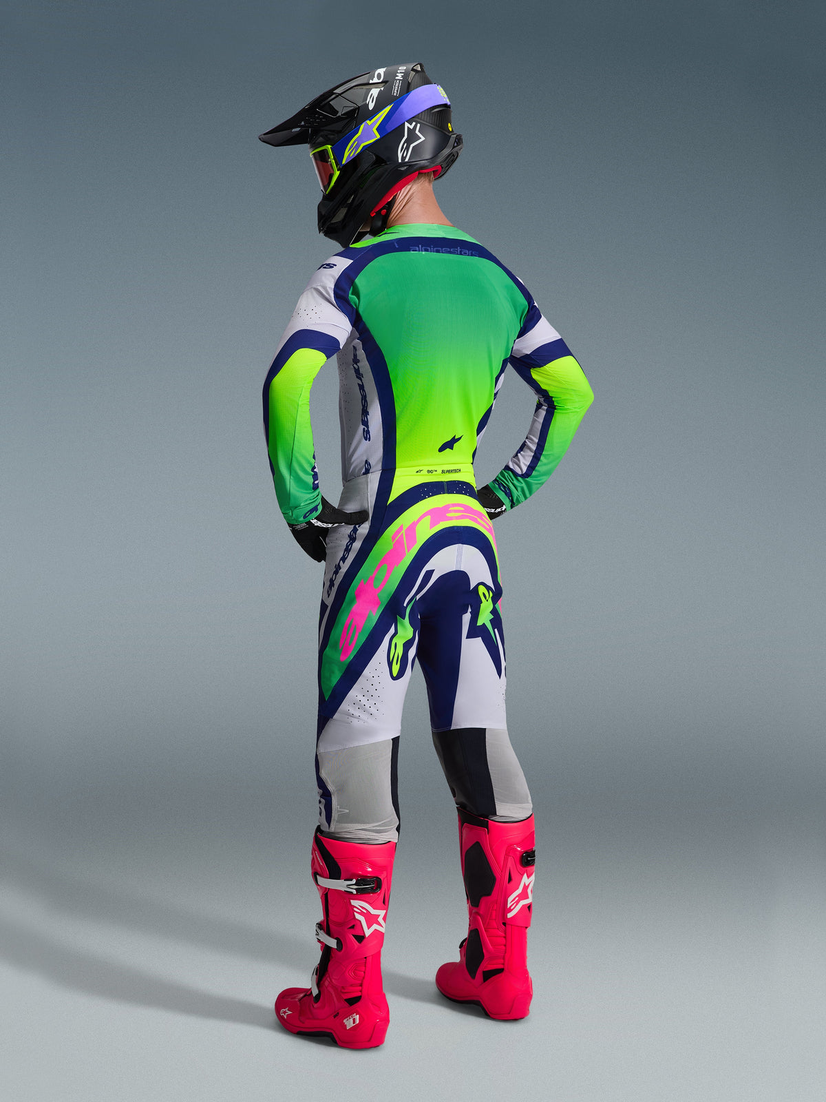 Zawodnik motocrossowy widziany od tyłu w koszulce Supertech Vista 2026 i pasujących spodniach w kolorach jasnoszarym, żółtym fluo i niebieskim, jaskraworóżowe buty i czarny kask, pełny zestaw wyścigowej odzieży off-road Alpinestars.