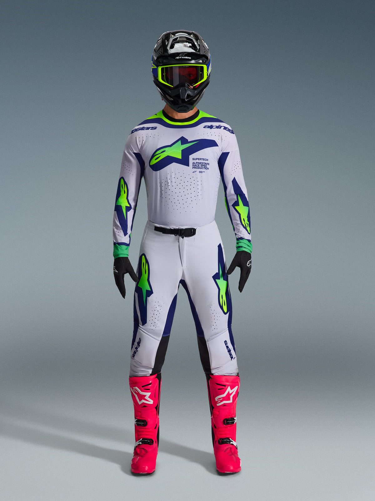 Zawodnik motocrossowy ubrany w koszulkę 2026 Supertech Vista w kolorze jasnoszarym, żółtym fluo i niebieskim, w komplecie z pasującymi spodniami, jaskraworóżowymi butami i czarnym kaskiem z neonowymi akcentami, profesjonalny sprzęt do wyścigów off-road.