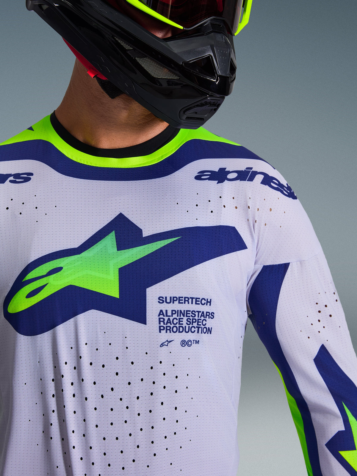 Alpinestars 2026 Supertech Vista Jersey, koszulka motocrossowa, jasnoszara z żółtymi fluo i niebieskimi akcentami, noszona przez motocyklistę w czarnym kasku i neonowo-żółtych goglach, posiada duże zielono-niebieskie logo Astars, laserowo wycinane otwory wentylacyjne, sportowy krój wyścigowy