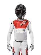 Motocyklista motocrossowy w koszulce Alpinestars 2024 Supertech Ward Jersey w kolorze White Hot Orange, biała koszulka z elastycznej siateczki z wyraźną czerwoną grafiką i logo na klatce piersiowej, czarny kask z ciemnymi goglami, białe rękawice, profesjonalny sprzęt do jazdy off-road