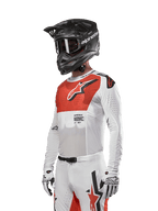 Alpinestars 2024 Supertech Ward Jersey, koszulka motocrossowa, biała i intensywnie pomarańczowa, z czerwonym panelem na klatce piersiowej i białym logo gwiazdy, długie rękawy z laserowo wycinanymi perforacjami, noszona z czarnym kaskiem i białymi rękawiczkami, sportowy krój wyścigowy