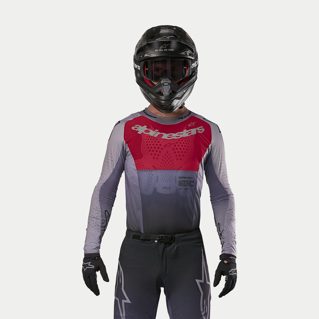 Motocyklista motocrossowy w koszulce Alpinestars 2024 Supertech Dade w kolorze Iron Red Berry, z czerwonym panelem na klatce piersiowej i szarymi rękawami ze srebrnym logo, w komplecie z pasującymi spodniami, czarnym kaskiem i czarnymi rękawicami, profesjonalny strój do wyścigów off-road