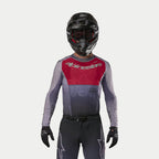 Motocyklista motocrossowy w koszulce Alpinestars 2024 Supertech Dade w kolorze Iron Red Berry, z czerwonym panelem na klatce piersiowej i szarymi rękawami ze srebrnym logo, w komplecie z pasującymi spodniami, czarnym kaskiem i czarnymi rękawicami, profesjonalny strój do wyścigów off-road
