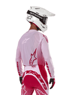 Motocyklista motocrossowy widziany od tyłu w koszulce Alpinestars 2024 Supertech Dade w kolorze Red Berry Lilac, z oddychającymi panelami z siatki i czerwonymi logo Alpinestars, w zestawie z pasującymi czerwono-liliowymi spodniami oraz białym kaskiem Alpinestars z goglami, profesjonalny sprzęt do wyścigów off-road