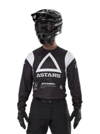 Alpinestars Techdura Jersey, czarna koszulka enduro, duża biała grafika z trójkątem i logo ASTARS na klatce piersiowej, białe panele na ramionach, motocyklista w czarnym kasku motocrossowym, goglach i pasującym czarnym stroju terenowym
