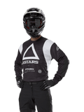 Alpinestars Techdura Jersey, koszulka motocrossowa, czarno-biała, noszona z pasującym kaskiem i rękawicami, poza z rękami na biodrach na białym tle, z dużym trójkątnym logo A i brandingiem ASTARS