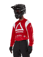 Alpinestars Techdura Jersey, koszulka motocrossowa, jaskrawoczerwona i biała, z wyraźnym wzorem białego trójkąta i brandingiem ASTARS, noszona z czarnym kaskiem i rękawicami, wytrzymała i odprowadzająca wilgoć odzież terenowa