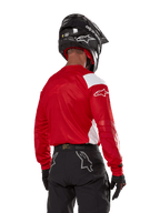 Motocyklista motocrossowy widziany od tyłu w koszulce Alpinestars Techdura w kolorze jasnoczerwonym z białymi panelami, zestawionej z czarnymi spodniami off-road, czarnym kaskiem i czarnymi rękawicami; dopasowany zestaw odzieży do jazdy enduro i motocrossowej.