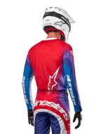 Zawodnik motocrossowy od tyłu w koszulce Alpinestars 2024 Techstar Pneuma w kolorze Blue Mars Red White, czerwony tył z białym logo gwiazdy, niebieskie i fioletowe rękawy z białym brandingiem, w komplecie z pasującymi spodniami i białym kaskiem do jazdy terenowej.