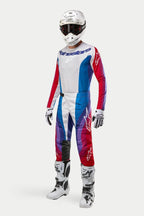 Kierowca motocrossowy w pełnym stroju Alpinestars, spodnie 2024 Techstar Pneuma w kolorze Blue Mars Red White, dopasowana koszulka z długim rękawem, biały kask z ciemnymi goglami oraz biało-czarne buty, kompletny zestaw do wyścigów off-road