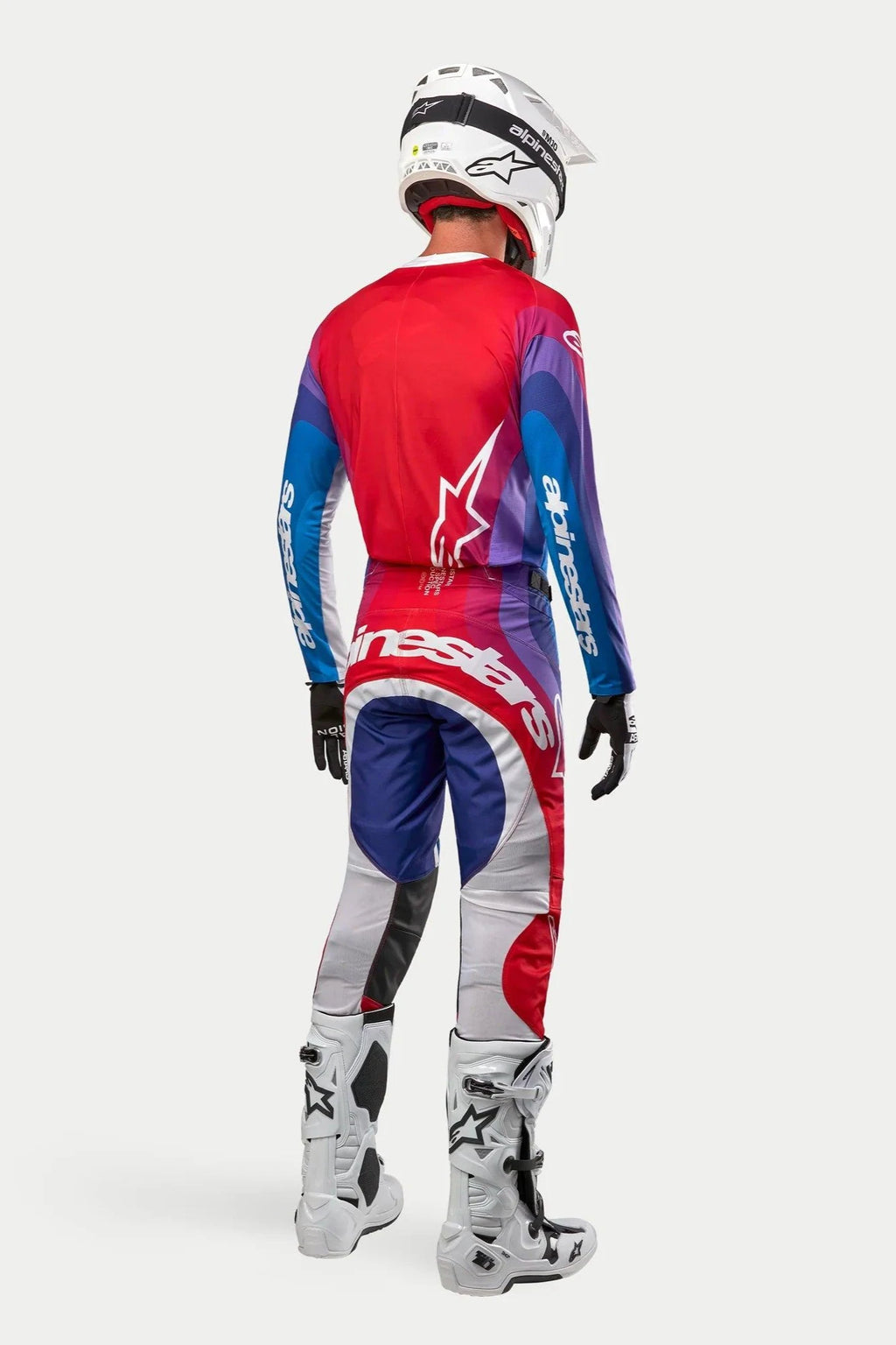 Zawodnik motocrossowy widoczny od tyłu w bluzie i spodniach Alpinestars 2024 Techstar Pneuma, kolorystyka Blu Mars Red White z czerwonymi, niebieskimi i fioletowymi gradientami, białe logo, białe buty i biały kask, dopasowany strój do wyścigów terenowych