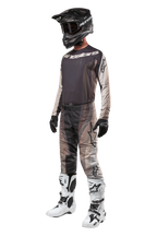Kierowca motocrossowy w pełnym stroju wyścigowym, koszulka 2024 Techstar Pneuma w kolorach Dark Sand i Iron Dust Gray, pasujące wzorzyste spodnie motocrossowe, czarny kask z goglami, biało-czarne buty, czarne rękawice, skoordynowany zestaw do wyścigów terenowych