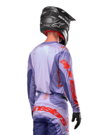 Alpinestars 2024 Techstar Ocuri Jersey, koszulka motocrossowa, fioletowa i jaskrawopomarańczowa, widok z tyłu na motocyklistę w pasującym komplecie i czarnym kasku, duże pomarańczowe napisy Alpinestars na rękawach i w dolnej części pleców