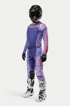 Zawodnik motocrossowy w koszulce Alpinestars 2024 Techstar Ocuri w kolorze fioletowo-pomarańczowym, pasujące fioletowe spodnie wyścigowe z pomarańczowymi akcentami, czarny kask, biało-czarne buty i czarne rękawice, kompletny profesjonalny strój off-road