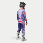 Strój motocrossowy Alpinestars Techstar Ocuri, fioletowy zestaw z jaskrawymi pomarańczowymi akcentami, widok od tyłu, koszulka i spodnie z logo Alpinestars, czarny kask, czarne rękawice, białe buty motocyklowe, sportowy design