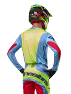 Motocyklista motocrossowy widoczny od tyłu w koszulce Alpinestars 2024 Techstar Ocuri, jasnoniebieska z neonowożółtym tyłem i czerwonymi akcentami na rękawach, dopasowane spodnie motocrossowe i neonowożółty kask, skoordynowany strój do wyścigów terenowych