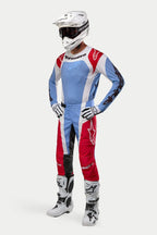 Zawodnik motocrossowy w pełnym stroju wyścigowym Alpinestars, koszulka 2024 Techstar Ocuri w kolorach jasnoniebieskim, czerwonym i białym, pasujące spodnie, biały kask z goglami, białe buty i biało-czarne rękawice