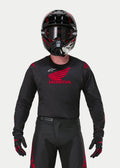 Motocyklista motocrossowy ubrany w koszulkę Alpinestars Honda Racer Iconic w kolorze czarno-czerwonym, z dużym czerwonym logo skrzydła Honda na piersi i białym logo Astar, w zestawie z dopasowanym czarnym kaskiem, goglami i rękawicami tworzącymi kompletny strój wyścigowy off-road