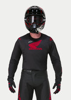 Motocyklista motocrossowy ubrany w koszulkę Alpinestars Honda Racer Iconic w kolorze czarno-czerwonym, z dużym czerwonym logo skrzydła Honda na piersi i białym logo Astar, w zestawie z dopasowanym czarnym kaskiem, goglami i rękawicami tworzącymi kompletny strój wyścigowy off-road