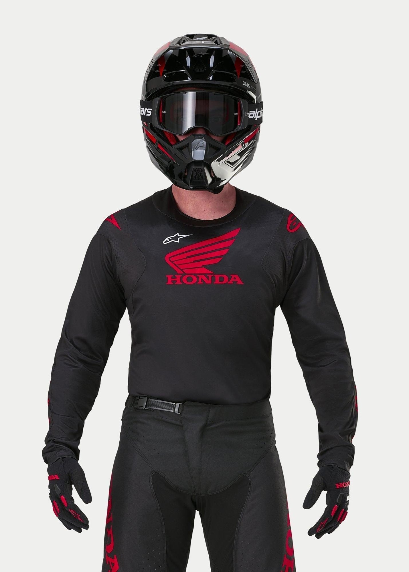 Motocyklista motocrossowy ubrany w koszulkę Alpinestars Honda Racer Iconic w kolorze czarno-czerwonym, z dużym czerwonym logo skrzydła Honda na piersi i białym logo Astar, w zestawie z dopasowanym czarnym kaskiem, goglami i rękawicami tworzącymi kompletny strój wyścigowy off-road