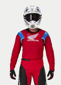 Motocyklista motocrossowy w koszulce Alpinestars Honda Racer Iconic, jaskrawoczerwona z niebieskimi i białymi akcentami oraz logo Hondy, biało-niebieski kask, ciemne gogle i pasujące czerwono-czarne spodnie motocrossowe, odzież wyczynowa do jazdy terenowej
