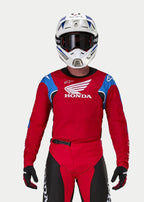Motocyklista motocrossowy w koszulce Alpinestars Honda Racer Iconic, jaskrawoczerwona z niebieskimi i białymi akcentami oraz logo Hondy, biało-niebieski kask, ciemne gogle i pasujące czerwono-czarne spodnie motocrossowe, odzież wyczynowa do jazdy terenowej