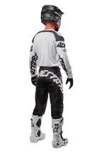Alpinestars 2024 Racer Hana Jersey, widok z tyłu na zawodnika motocrossowego, biało-czarna, biała koszulka z czarnymi grafikami na rękawach, czarne spodnie z białym napisem Alpinestars na siedzeniu, czarny kask oraz biało-czarne buty, dopasowany strój do wyścigów terenowych