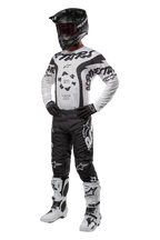 Zawodnik motocrossowy w pełnym rynsztunku, koszulka Alpinestars 2024 Racer Hana w kolorze biało-czarnym, czarne spodnie motocrossowe z białymi logo, czarny kask, biało-czarne buty wyścigowe i czarne rękawice, profesjonalny strój do wyścigów terenowych